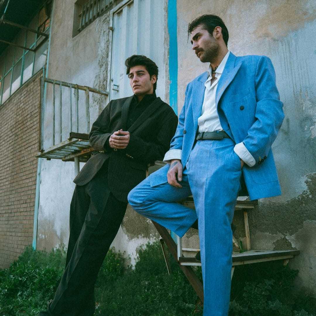 Two men in stylish suits pose confidently outside a rustic building.