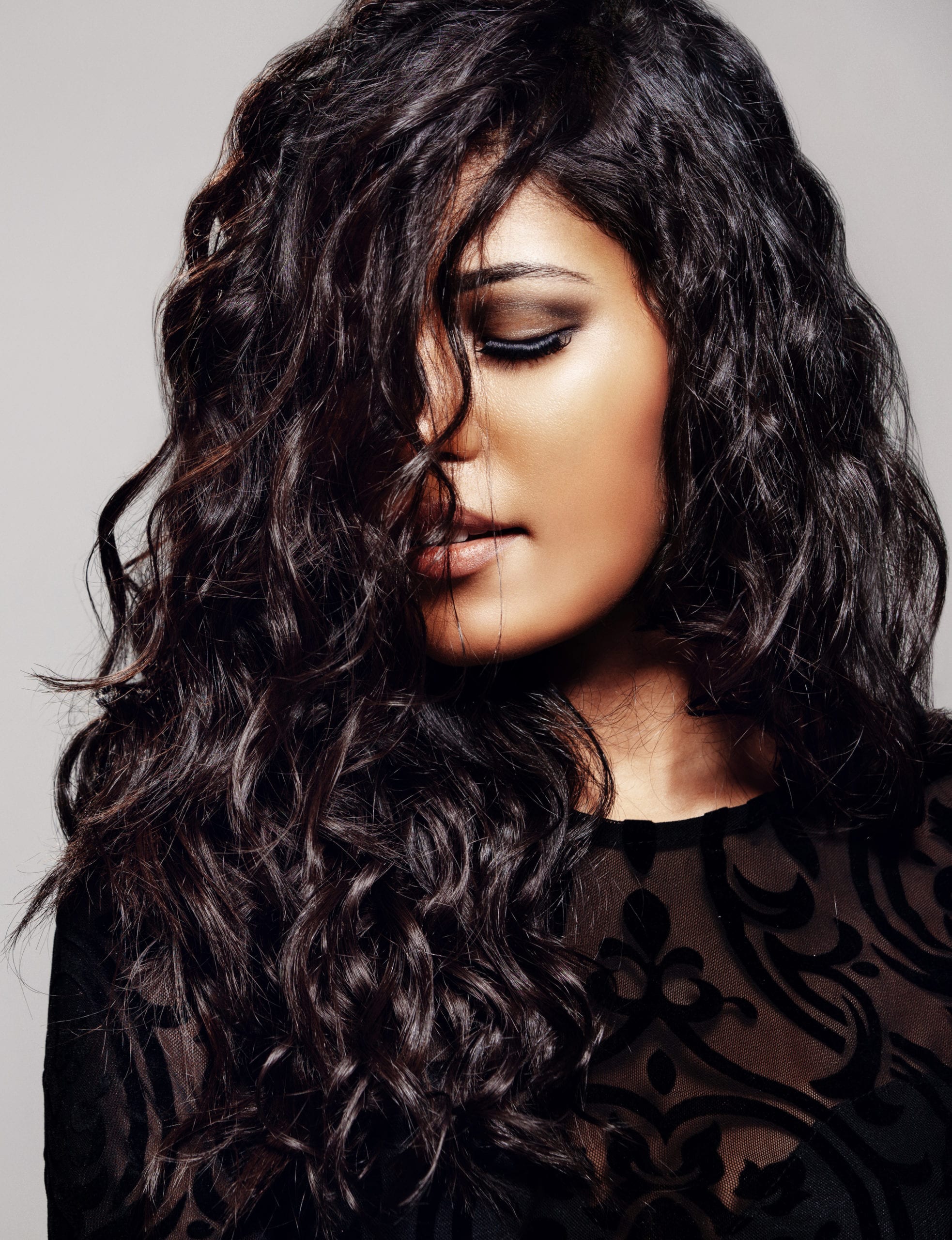 Sensual woman with shiny curly hair Sensual young woman with shiny curly hair. Female model with wavy hairstyle and eyes closed against grey background.